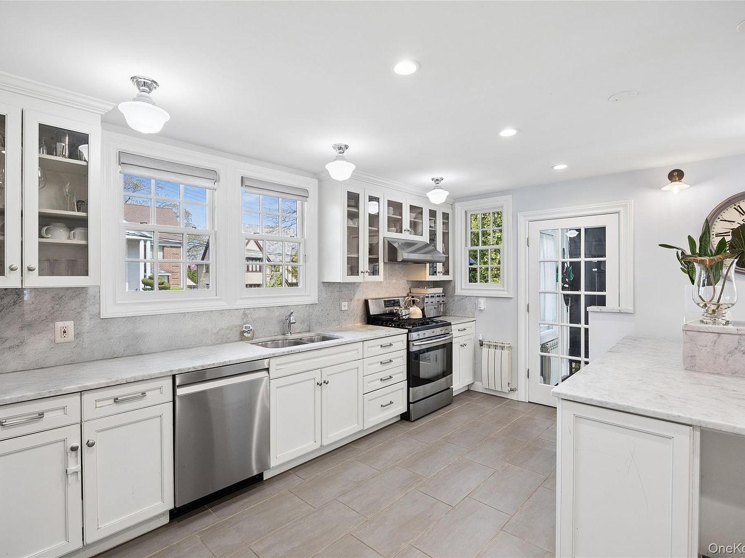 Renovated white kitchen with marble countertops, schoolhouse pendant lighting, stainless appliances and the original divided-light windows looking onto a Douglaston Manor streetscape of Tudor and Colonial Revival houses.